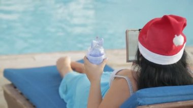 Havuzun kenarında Noel Baba şapkası takmış bir kadın video bağlantısı ve mutlu yıllar video bağlantısıyla akıllı telefonla tebrik ediyor. Arkadan ateş ediyorlar..