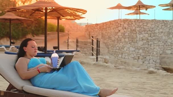 Женщина лежит в шезлонге на берегу моря, пьет коктейль и работает с ноутбуком, стреляя сбоку. — стоковое видео