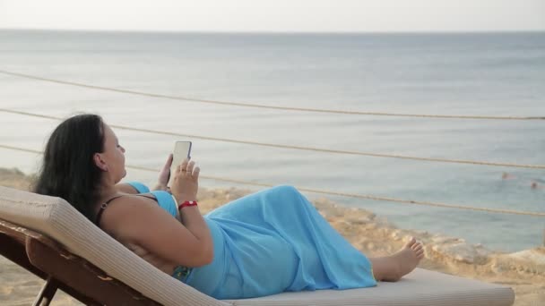 Una donna in un pareo in un lettino in riva al mare sta messaggiando in uno smartphone guardando il mare — Video Stock