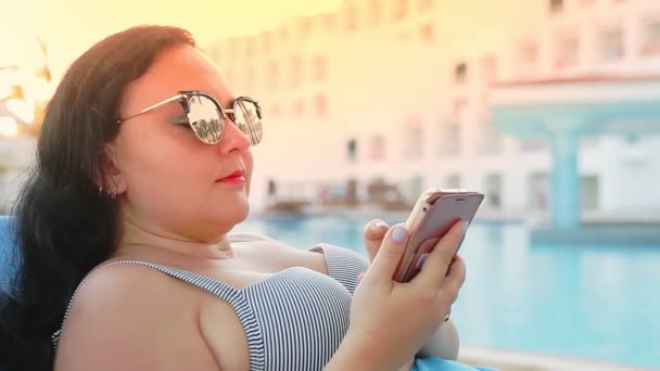 Брюнетка в купальнику, парео і сонцезахисні окуляри на шезлонгу біля басейну спілкується в смартфоні. Сонячне світло — стокове відео