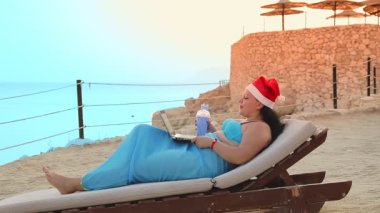 Deniz kıyısında Noel Baba şapkalı esmer bir kadın kokteyl içer ve dizüstü bilgisayarda parmak izi bırakır..
