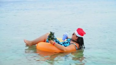 Noel Baba şapkalı ve güneş gözlüklü esmer bir kadın Noel tatilinde elinde bir Noel ağacıyla denizde yüzer ve kokteyl içer.