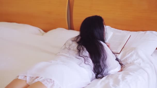 大きな広いベッドの上の白いローブの女性は彼女の胃の上に横たわる本を読んでいる — ストック動画