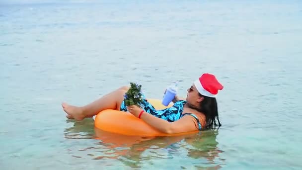 Egy barna nő télapó sapkában és napszemüvegben a karácsonyi ünnepek alatt körbeúszik a tengerben egy karácsonyfával a kezében és iszik egy koktélt. — Stock videók