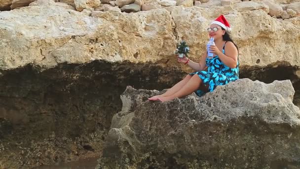 Brunetka v klobouku Santa Claus a slunečních brýlích sedí na skále dívá na moře a pije koktejl o vánočních svátcích — Stock video