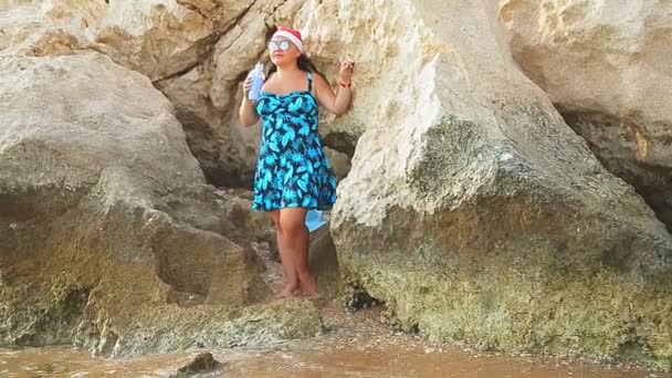 Noel Baba şapkalı esmer bir kadın deniz kıyısında duruyor ve Noel tatili boyunca kokteyl içiyor. — Stok video