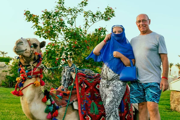 Family couple tourists next to the decorated lying camel woman with closed cape face. — Stock Photo, Image