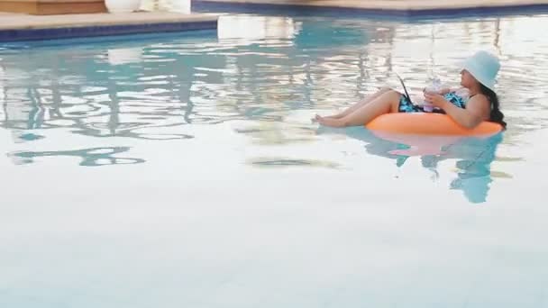 Μια γυναίκα με καπέλο ήλιου και ένα μαγιό στην πισίνα σε ένα δαχτυλίδι κολύμβησης κολυμπά αργά και αναπαύεται στην περιοχή του ξενοδοχείου — Αρχείο Βίντεο