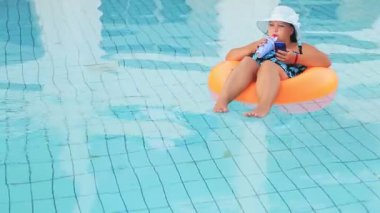 Güneş şapkalı Yahudi kadın ve havuzda mayo, akıllı telefonlu bir yüzme çemberinde kokteyl içiyor..