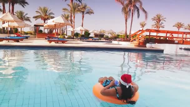 Havuzda yüzme çemberinde Noel Baba şapkalı bir kadın, arka planda palmiye ağaçları ve güneşli yerlerde kameraya arkası dönük bir şekilde kokteyl içiyor. — Stok video