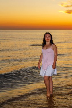 Beyaz elbiseli esmer bir kadın gün batımında deniz kıyısında duruyor..