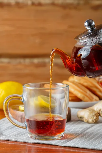 Сильный насыщенный чай наливают в чашку из стеклянного чайника рядом с лимоном и имбирем. — стоковое фото