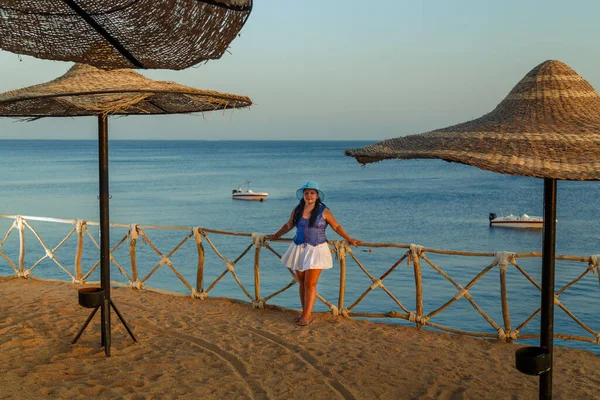Uma jovem mulher em uma saia branca e um chapéu de sol no fundo do mar admira a natureza da costa. — Fotografia de Stock
