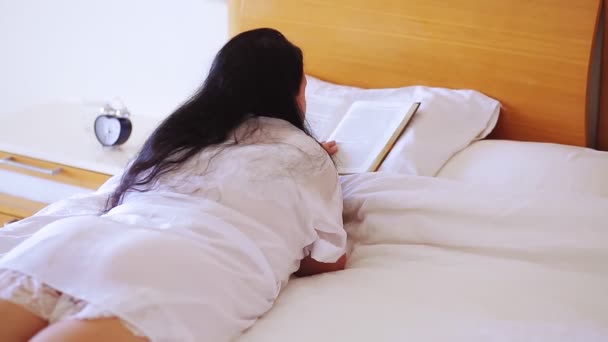 Uma mulher em um manto branco em uma grande cama larga está lendo um livro deitado em seu estômago — Vídeo de Stock