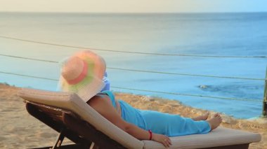 Şapkalı bir kadın, deniz kıyısında güneşlenir ve arkasından denize bakar.