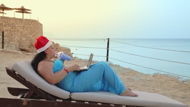 Μια γυναίκα στην παραλία σε μια ξαπλώστρα με μαγιό παρεό και ένα καπέλο του Άι Βασίλη πίνει ένα κοκτέιλ. — Αρχείο Βίντεο