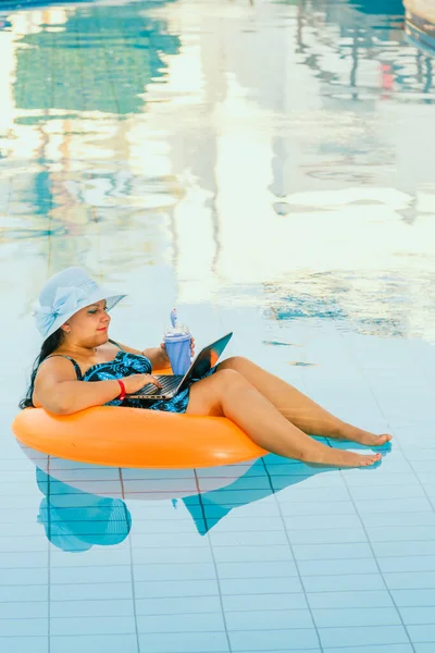 Havuzda güneş şapkalı bir kadın kokteyl ve dizüstü bilgisayarla video bağlantısıyla iletişim kuruyor. Dikey fotoğraf