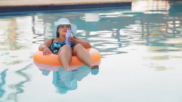 Donna gioiosa in un cappello da sole e un costume da bagno in piscina in un cerchio di nuoto nuota lentamente e beve una bibita analcolica e riposa — Video Stock