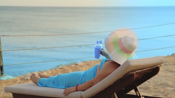Kobieta w kapeluszu leeit w leżaku na brzegu morza i patrzy na morze od tyłu — Wideo stockowe