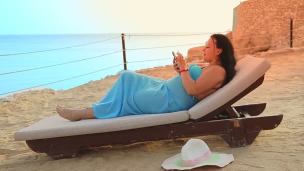Una donna si trova in un lettino sulla riva del mare a bere un cocktail e sta chattando in uno smartphone dal lato. — Video Stock