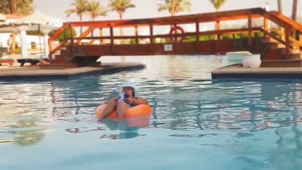 Morena mujer en un sombrero de sol y traje de baño en la piscina en el círculo de natación con un teléfono inteligente bebe un cóctel. — Vídeos de Stock