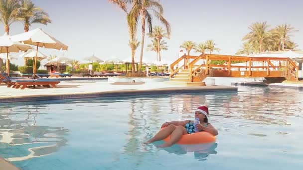 Een vrouw in een zwemcirkel in het zwembad in een kerstmuts drinkt een cocktail op vakantie tegen de achtergrond van palmbomen en ligstoelen — Stockvideo