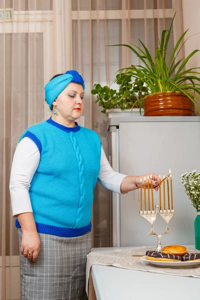 Uma mulher judia em uma cobertura para a cabeça na sala de jantar luzes de cozinha velas em Hanukkah na mesa ao lado de donuts e um vaso de flores. — Fotografia de Stock