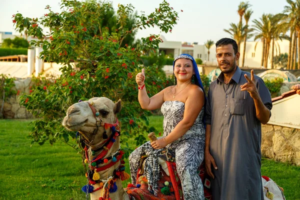 Egypt, Sharm El Sheikh, August 30, 2021 a woman in a cape on her head next to a Bedouin and a camel and lying on the grass on a city street. — Stock Photo, Image