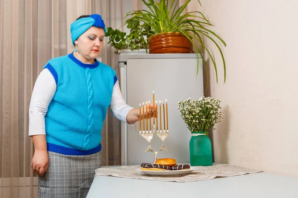 一名犹太妇女头戴头巾，在光明节的桌上点燃蜡烛，旁边放着甜甜圈和一个花瓶. — 图库照片