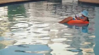 Yüzme havuzunda güneş şapkalı ve mayo giymiş bir kadın otelin bölgesinde yavaşça yüzer ve dinlenir..