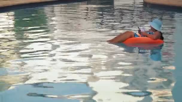Een vrouw met een zonnehoed en een badpak in het zwembad in een zwemring zwemt langzaam en rust op het grondgebied van het hotel. — Stockvideo