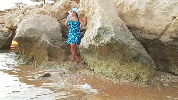 Una donna bruna con cappello e occhiali da sole si trova sulla riva rocciosa del mare e beve un cocktail guarda il mare. — Video Stock