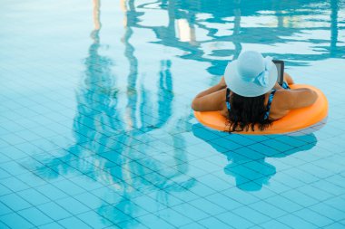 Yüzme havuzunda güneş şapkalı bir kadın elinde dizüstü bilgisayarla arkadan ateş ederken gevşiyor..
