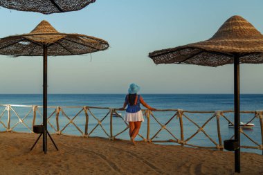 Beyaz etekli, güneş şapkalı, arka planda duran genç bir kadın sırtı ve gökyüzüne ve denize bakıyor..
