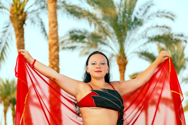 En brunett kvinna i en röd orientalisk dansdräkt på en bakgrund av palmer dansar med en mantel. — Stockfoto