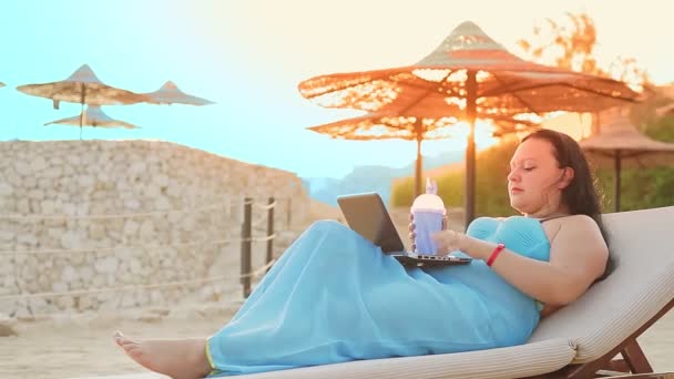 Μια γυναίκα σε μια ξαπλώστρα στην παραλία πίνει ένα κοκτέιλ και μιλάει με ένα gadget — Αρχείο Βίντεο