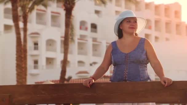 Брюнетка еврейка в синей шляпе стоит на мосту, любуясь пальмами и бассейном на фоне отеля — стоковое видео
