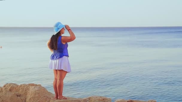 一个戴着蓝色帽子的黑发女人背对着摄像机站在岩石上欣赏大海和地平线. — 图库视频影像