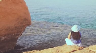 Mavi şapkalı esmer bir kadın sırtı taşların üzerinde oturur ve denize hayranlıkla bakar.