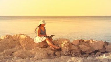 Mavi şapkalı esmer bir kadın, kayalık bir deniz kıyısında, güneş ışınlarında uzaktan bilgisayarla çalışıyor..