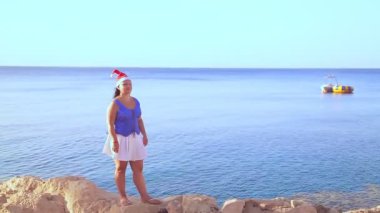 Noel Baba şapkalı esmer bir kadın kayalık sahil boyunca yürür ve doğaya hayran kalır.