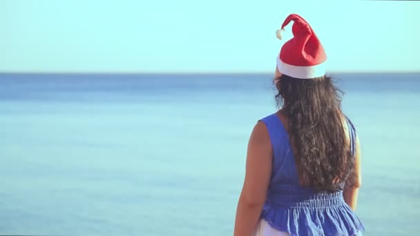 Una mujer morena en un sombrero de Santa Claus está de pie en la orilla del mar con su espalda a la cámara — Vídeo de stock