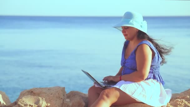 Une femme brune coiffée d'un chapeau bleu sur un littoral rocheux travaille à distance avec un ordinateur portable — Video