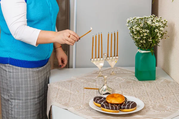 Еврейка в столовой зажигает свечи на Хануке на столе рядом с пончиками и вазой с цветами. — стоковое фото