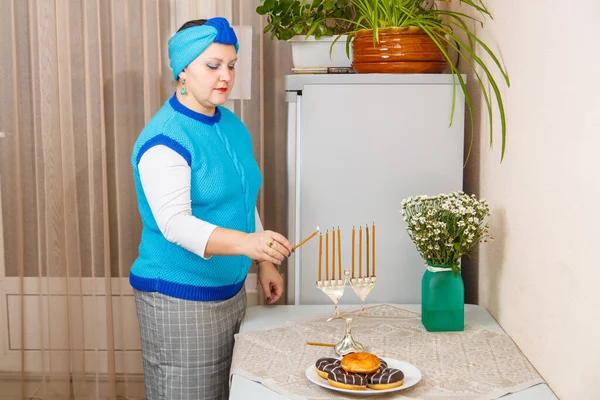 Una donna ebrea con un giubbotto blu e un turbante nella sala da pranzo accende candele a Hanukkah sul tavolo accanto a ciambelle e un vaso di fiori.. — Foto Stock