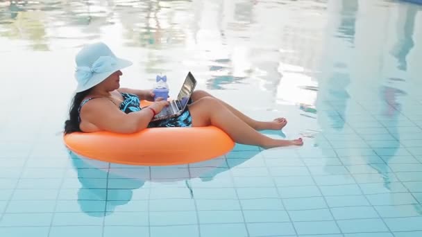 Mujer alegre en un sombrero de sol y un traje de baño en la piscina en el círculo de natación bebe un cóctel y descansa — Vídeos de Stock