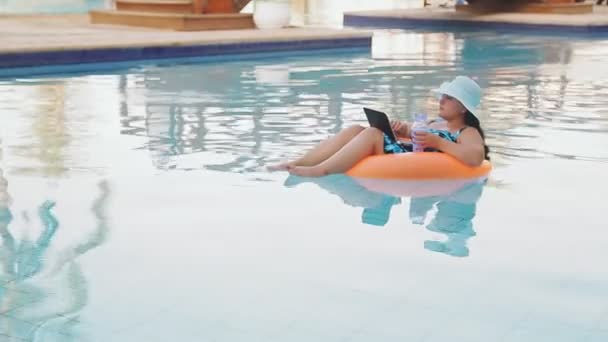 Donna in un cappello e costume da bagno in piscina in un cerchio di nuoto lentamente nuota con un computer portatile in grembo — Video Stock