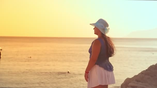 一个黑发女人，戴着蓝色的帽子，站在岩石海岸上，夕阳西下时闪烁着光芒，她欣赏大海和夕阳 — 图库视频影像