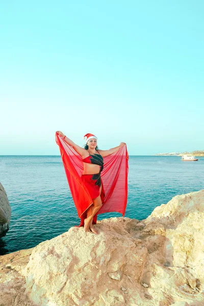 Μια γυναίκα με ανατολίτικη στολή χορού με καπέλο Αϊ Βασίλη χορεύει με ένα σάλι σε ένα βράχο δίπλα στη θάλασσα.. — Φωτογραφία Αρχείου