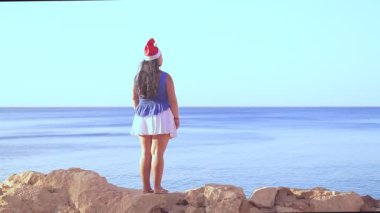 Noel Baba şapkalı esmer bir kadın, kayalık sahil boyunca yürür ve deniz yüzeyine hayran kalır.